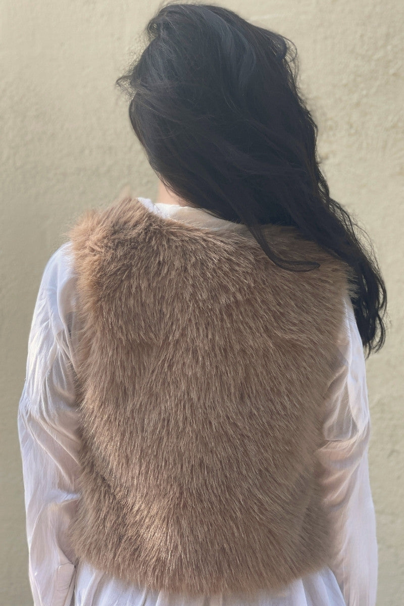 Back of woman with long dark hair wearing a taupe faux-fur vest over a white shirt against a plain background