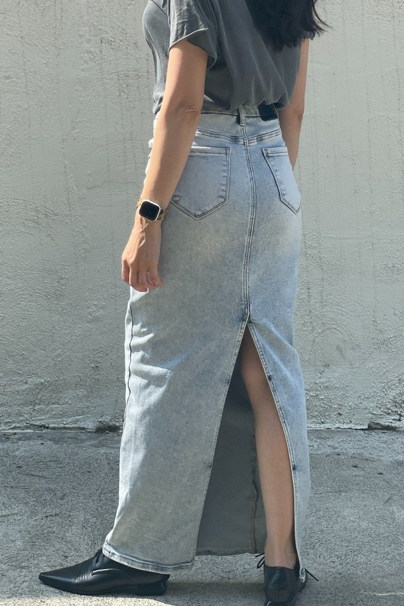 Person wearing a light blue denim maxi skirt with a high slit, standing against a plain wall.