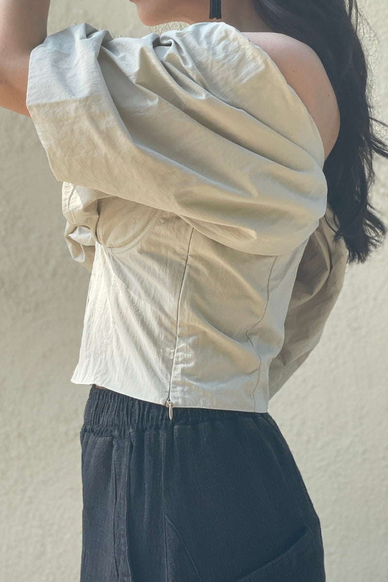 Woman with dark hair, standing sideways wearing a beige off-shoulder top and dark pants against a neutral background