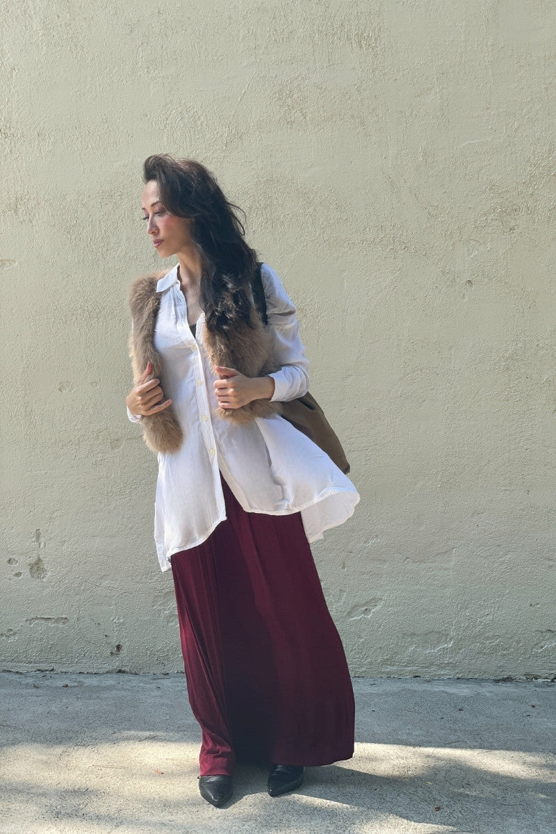Woman wearing a white blouse, taupe fur vest, and maroon skirt against a beige wall.