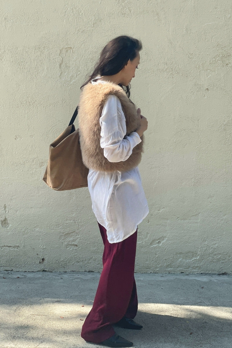 Dark haired woman wearing a faux-fur vest and holding a brown suede bag against a plain wall