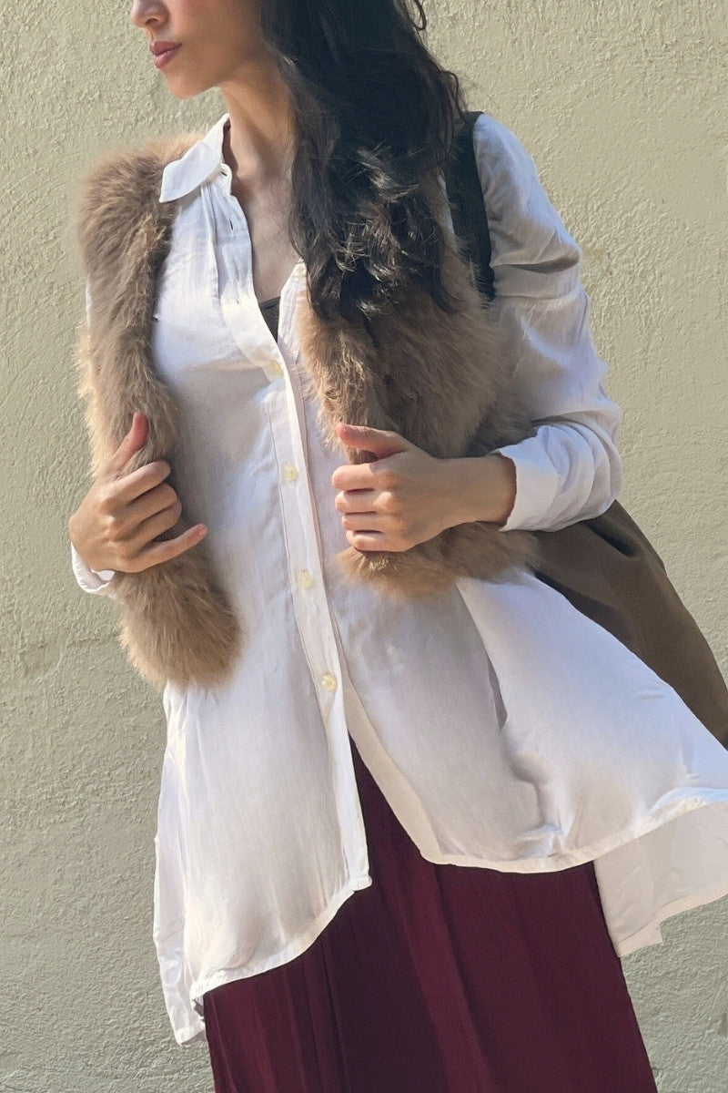 Woman wearing a white shirt and maroon skirt with a faux-fur vest against a beige wall.