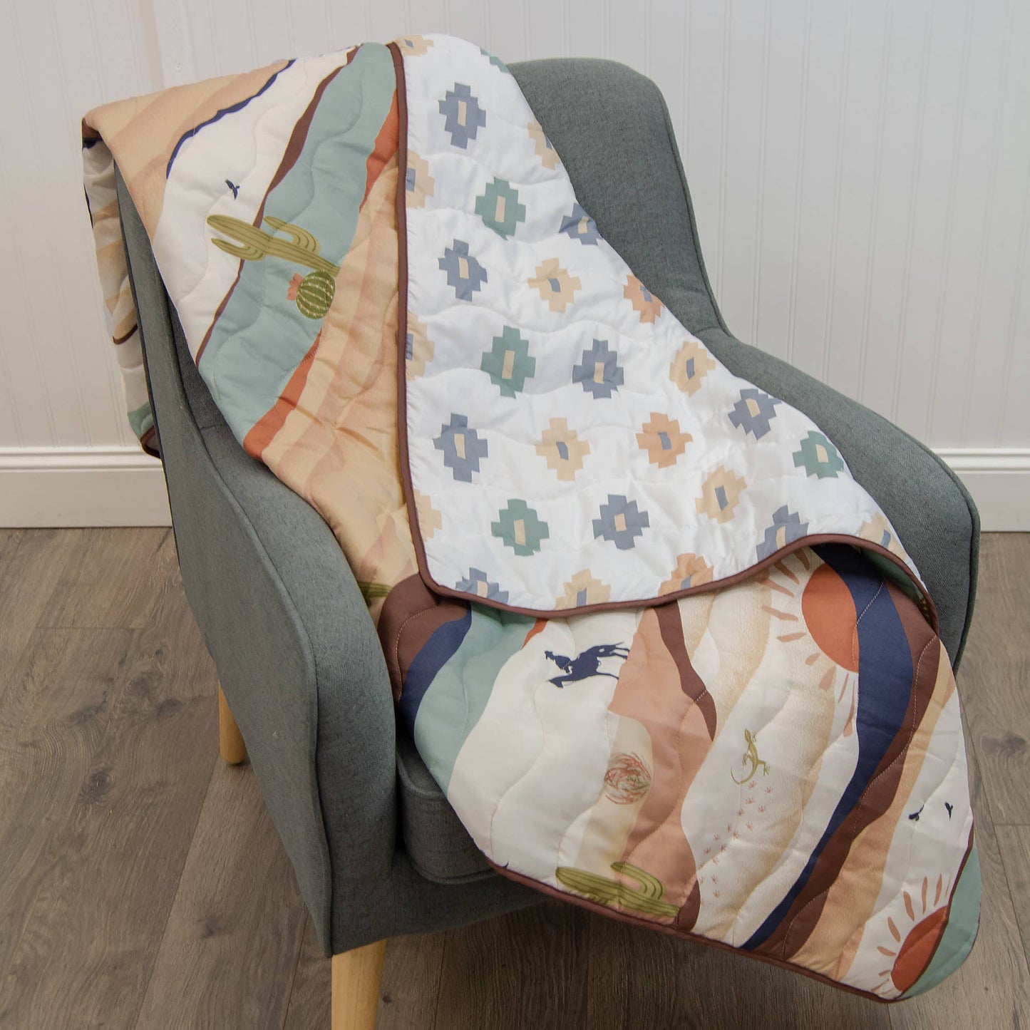 Earth-toned woven Journey throw with cowboy, cactus and sun design, draped over a neutral chair.