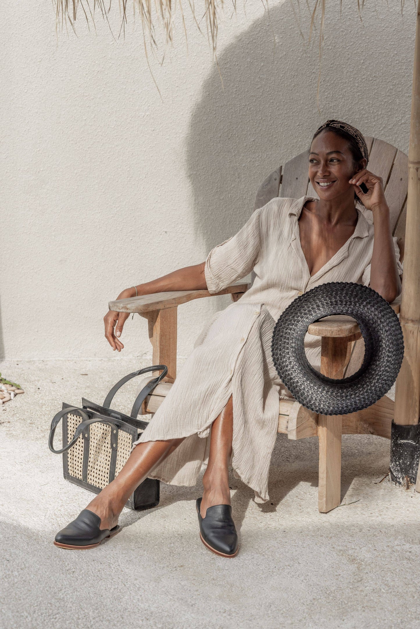 Model sits on wooden chair in sun with Papale Sun Hat in Black resting on the arm. Styled with beige linen dress and black mules.