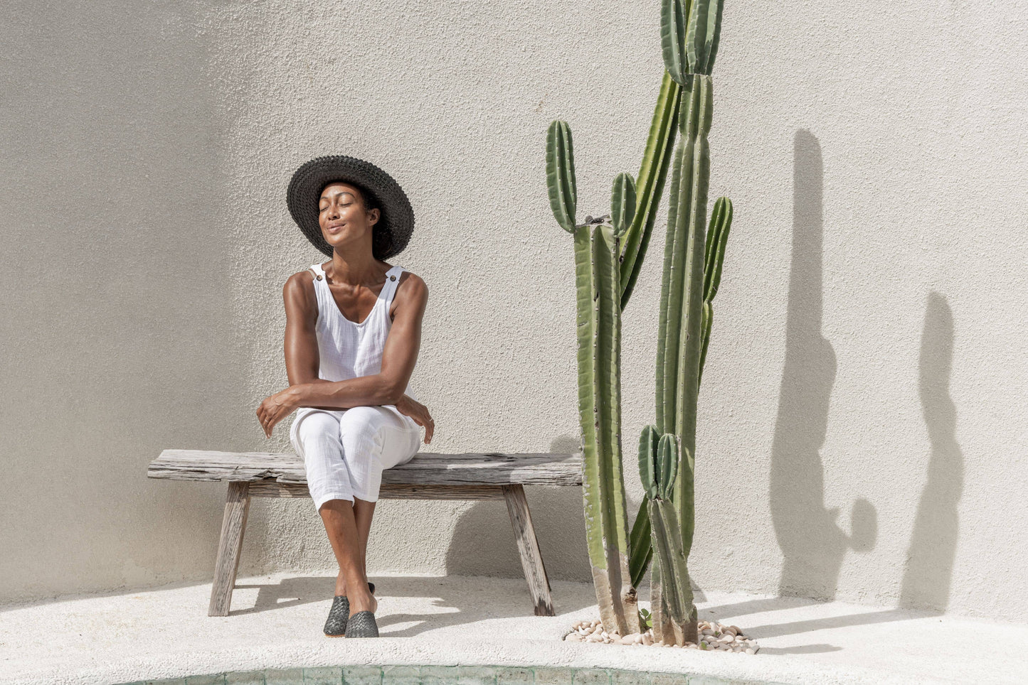 Lifestyle photo of Papale Sun Hat in Black. Model sits on wooden bench, beside cactus basking in the sun. Styled with white overalls.