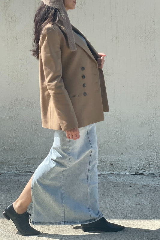 Woman wearing a beige blazer coat and light blue maxi skirt and bonnet against a plain background.