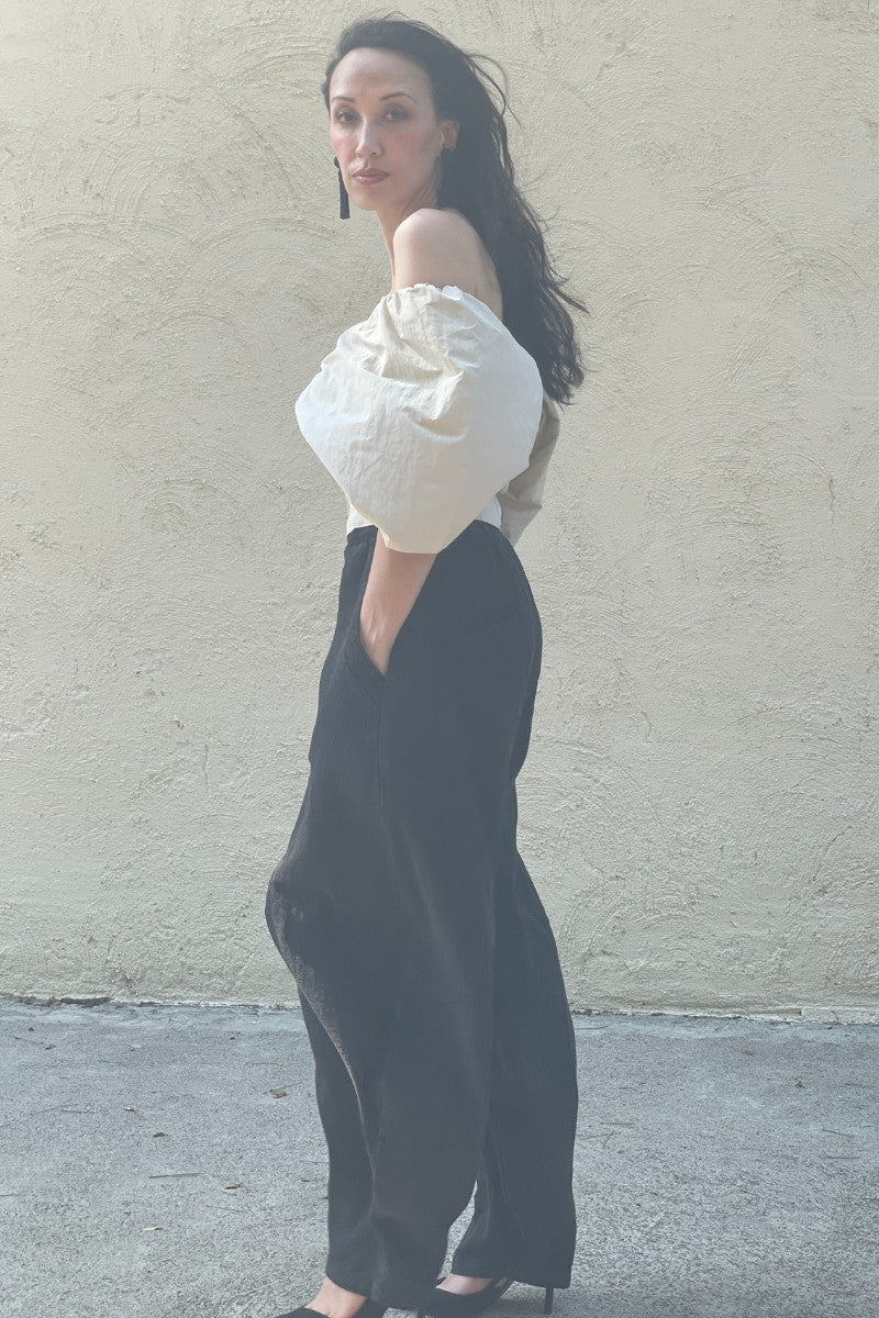 Woman wearing a beige off-shoulder top and black pants against a beige wall. Also wears black fringe earrings.