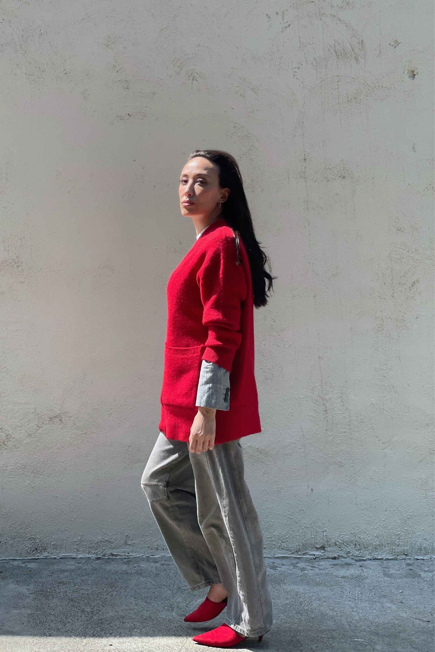 Model wearing a red cardigan, gray barrel jeans, and red mules standing against a plain wall.
