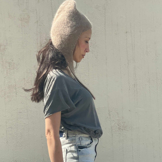 Woman wearing a beige knit hat and gray shirt against a beige wall. 
