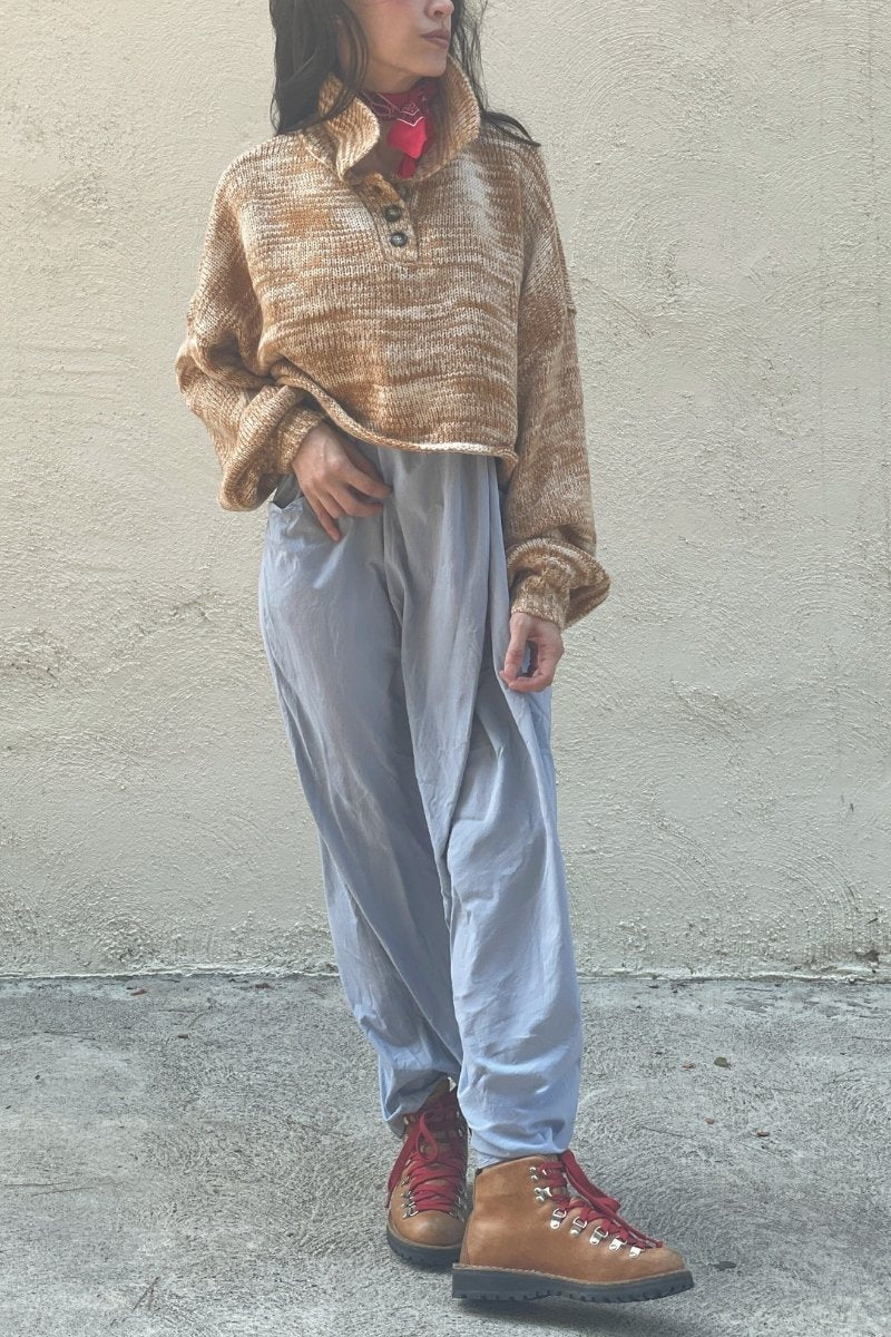 Woman wearing a rust colored sweater, with collar over a light blue dress, with orange hiking boots, and red laces, standing against a plain wall.