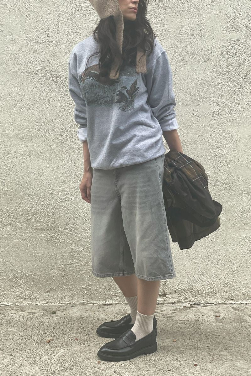 Woman wearing a gray vintage duck sweatshirt, grey denim jorts, beige socks and black loafers against a plain wall. Carrying a rain-jacket. 