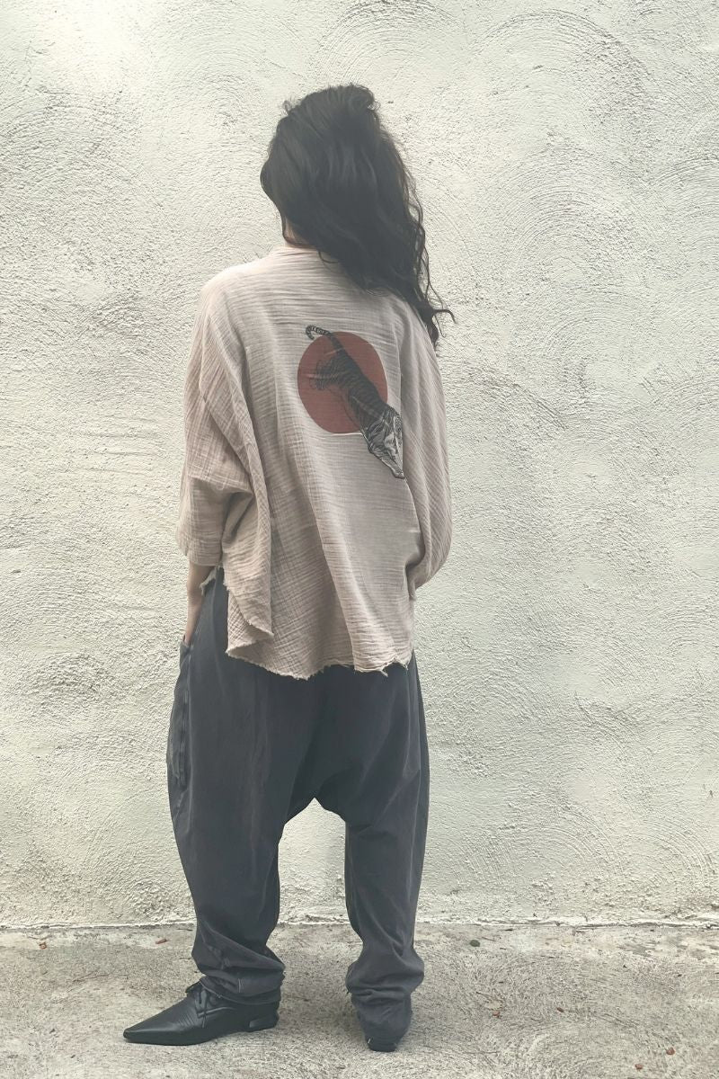 Model wearing a beige kimono with a sun and tiger design on back, facing  against a textured wall.