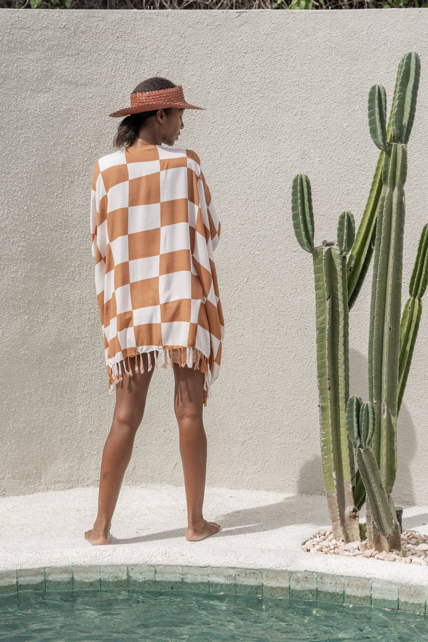 Back view of Papale Sun Hat in Clay. model stands by pool and cactus wearing hat and kimono. 