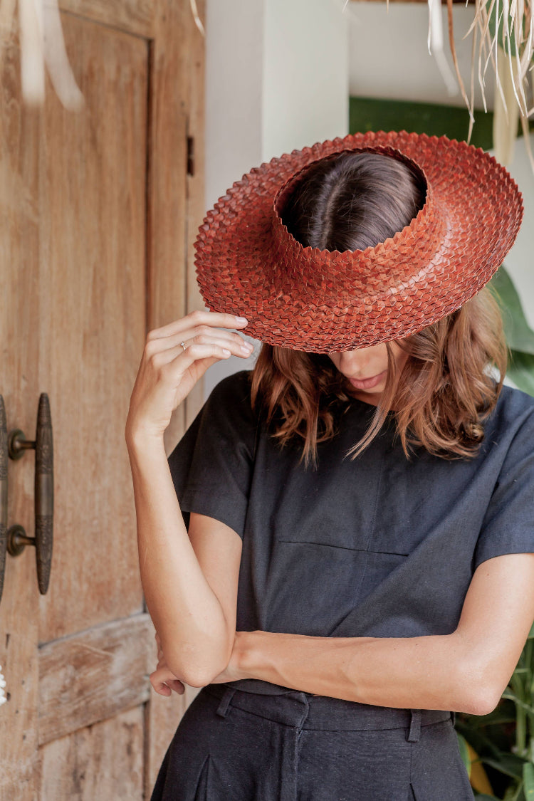 Handwoven dark clay-toned sun hat made from pandanus leaf, styled with black outfit outdoors.
