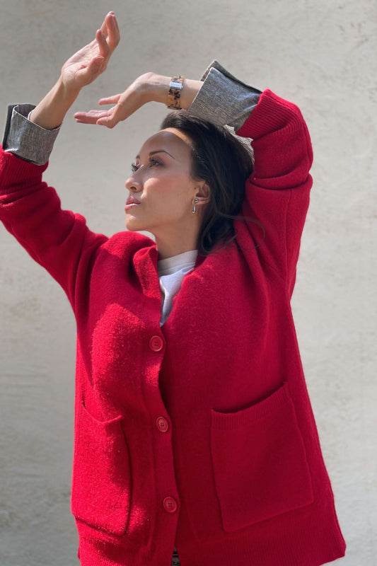 Model wearing a red cardigan, standing next to a white wall with hands in the air, blocking sunlight.