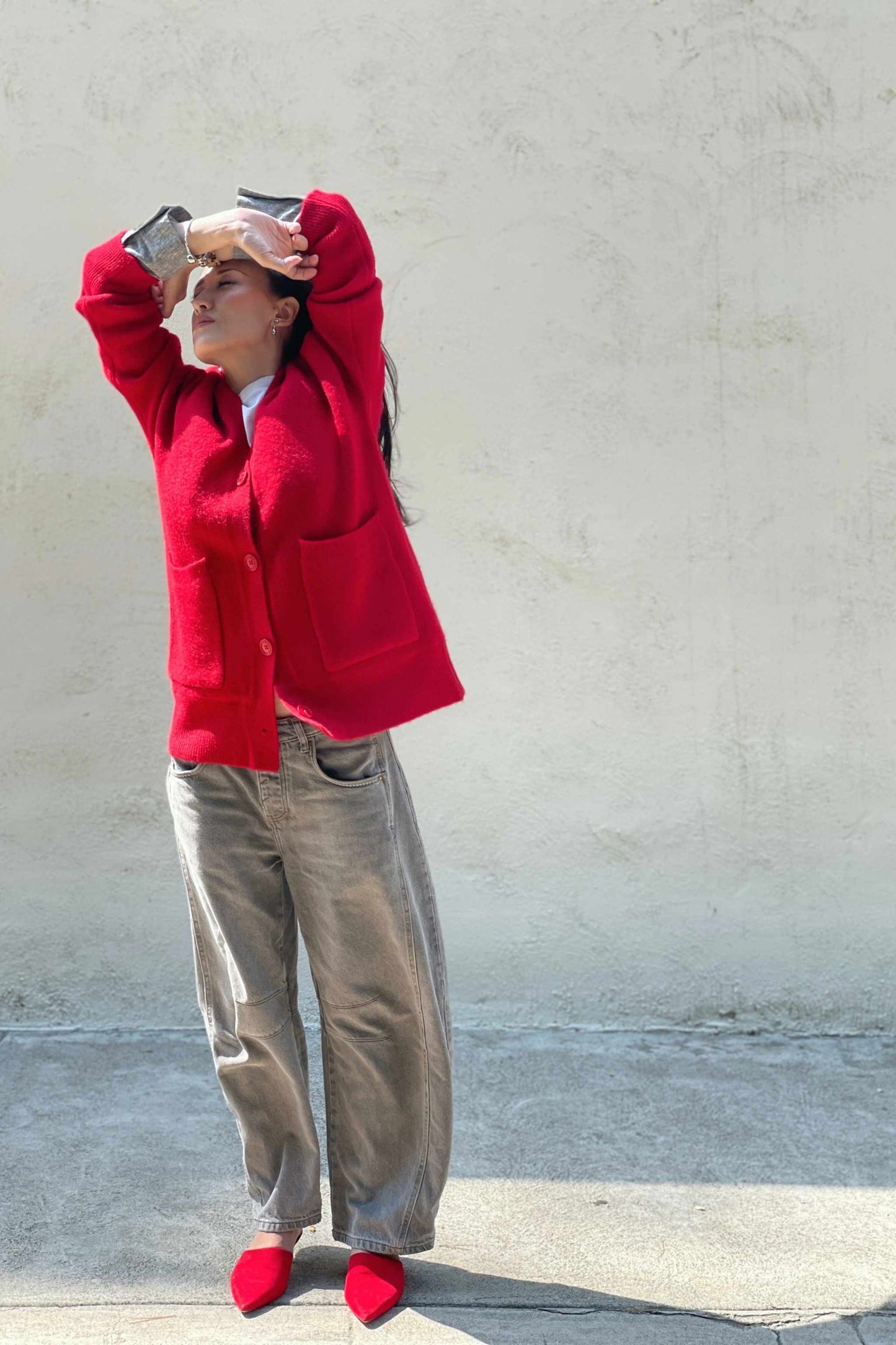 Model wearing a red cardigan, gray barrel jeans, and red mules, standing next to a white wall with hands in the air.