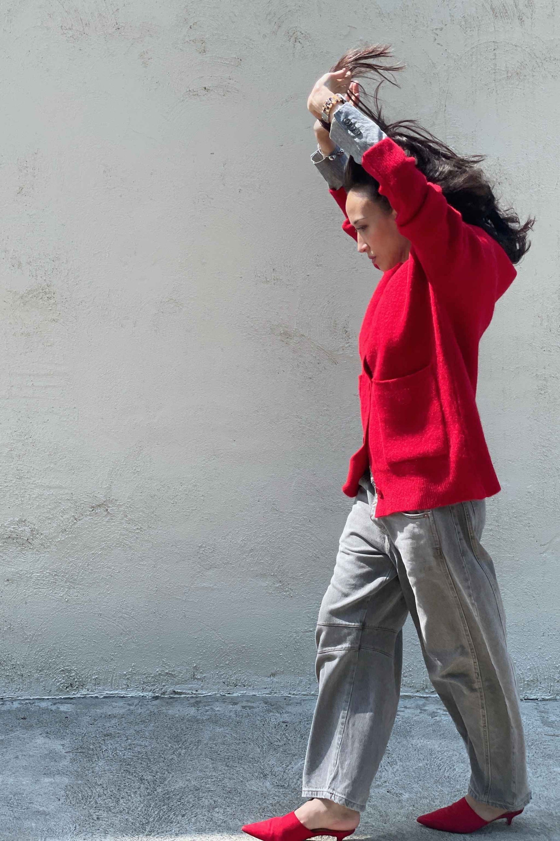 Model wearing a red cardigan, gray barrel jeans, and red mules, walking past a white wall with hands high in the air.