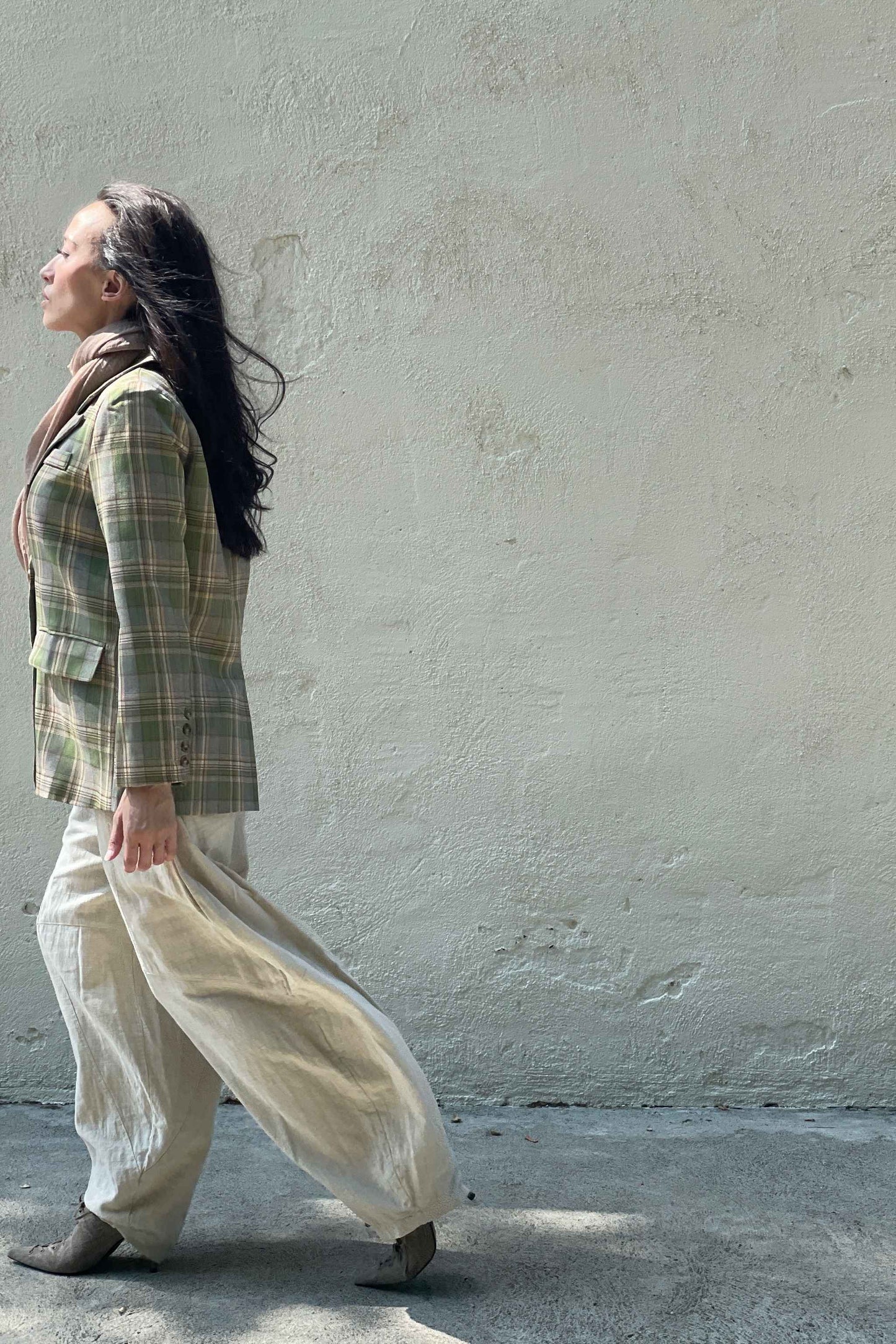 Model wearing Casual-Tartan-Color-Block-Blazer-in-Green near a neutral wall with sunlight on face, walking to the left. 