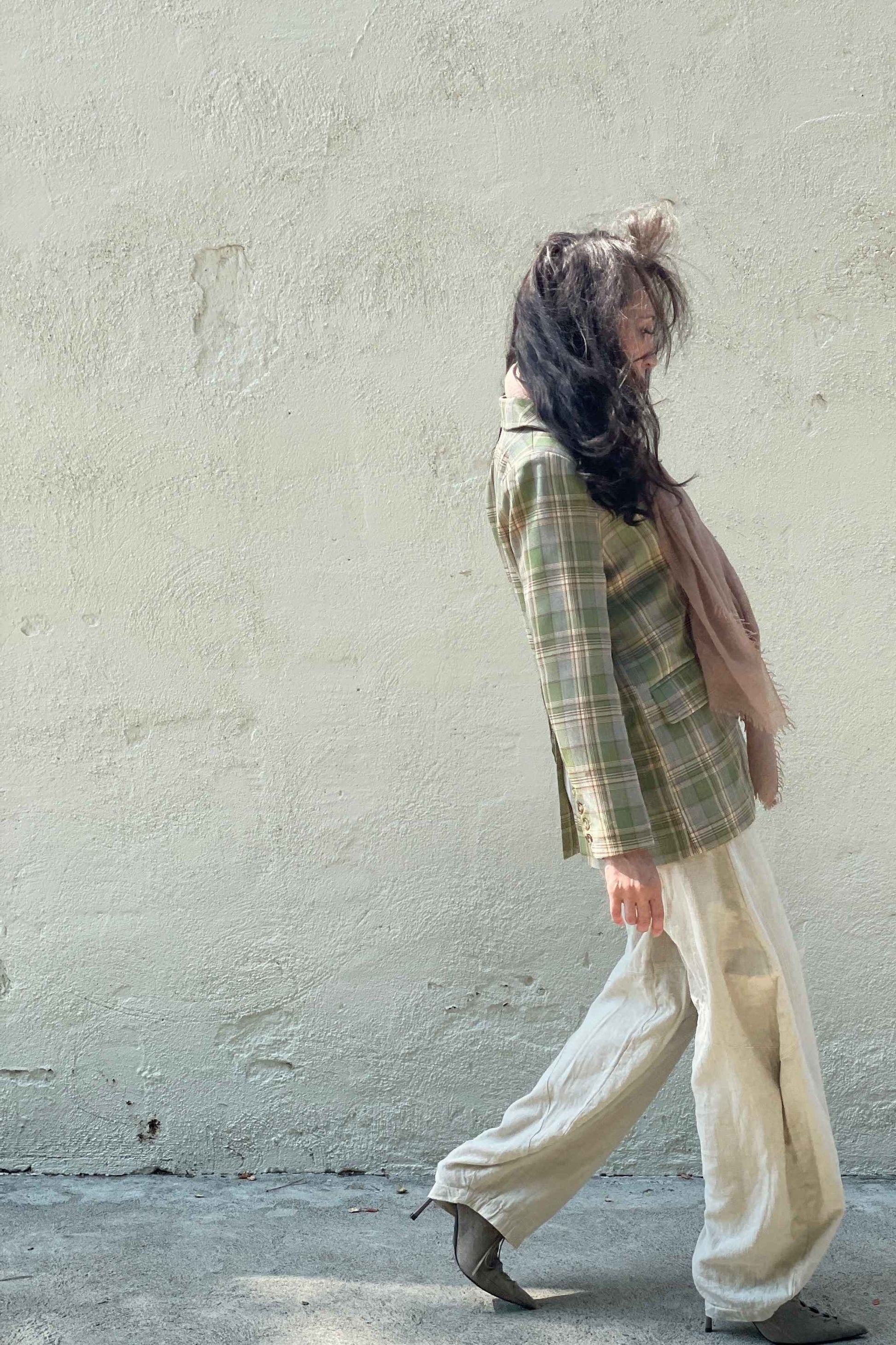 Model wearing Casual-Tartan-Color-Block-Blazer-in-Green near a neutral wall with sunlight on face, walking to the right. 