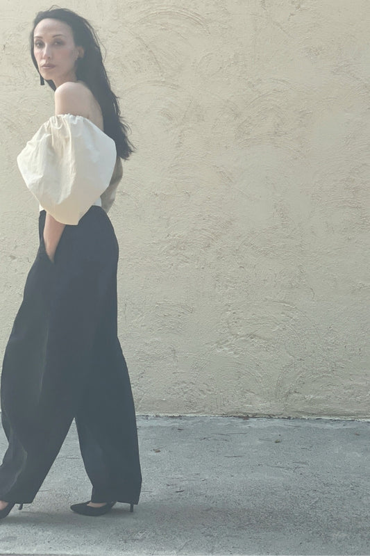 Model wearing a beige off-shoulder blouse and black pants against a beige wall. Exiting the frame.