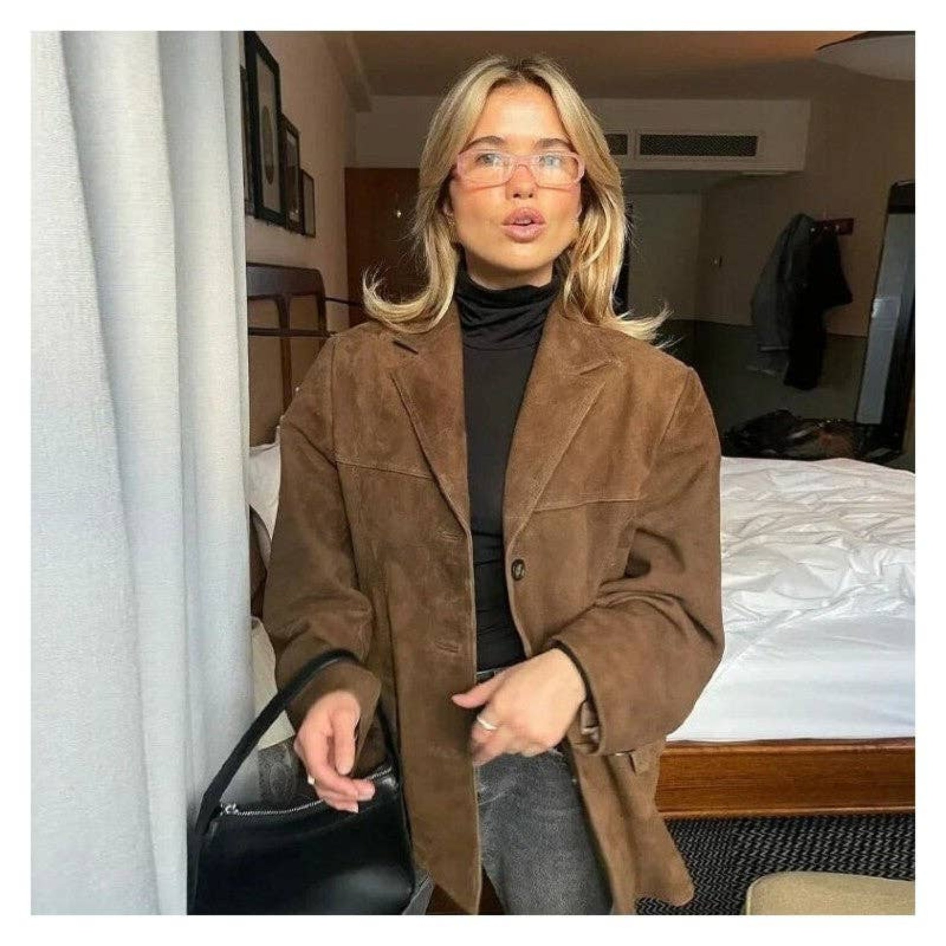 Brown cropped suede jacket with a minimal silhouette and vintage-inspired look. Model wears with glasses and black leather bag. 